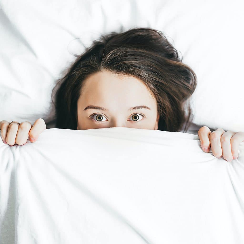 Wide-eyed woman in bed looking over a sheet trying to sleep better
